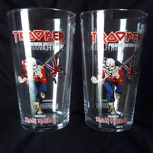 Puede incluir: Dos vasos de pinta de vidrio transparente con un gr&aacute;fico rojo y blanco de un hombre sosteniendo una bandera brit&aacute;nica. El texto "Trooper" est&aacute; impreso en rojo en el vaso. El texto "Premium British Beer" est&aacute; impreso en negro en el vaso. El texto "Created by Iron Maiden" est&aacute; impreso en rojo en el vaso.