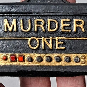 May include: A black and gold decorative box with the words "MURDER ONE" in gold lettering. The box has a series of red and gold buttons and a gold wreath with a star on each side. The box is held by a person.