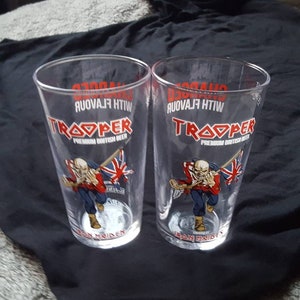 Puede incluir: Dos vasos de pinta de vidrio transparente con el logotipo de la cerveza Iron Maiden Trooper. El logotipo presenta una bandera de la Uni&oacute;n Jack roja, blanca y azul y el texto "Trooper Premium British Beer" con las palabras "With Flavour" impresas al rev&eacute;s.