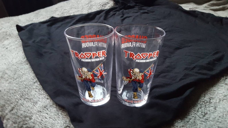 Puede incluir: Dos vasos de pinta de vidrio transparente con el logotipo de la cerveza Iron Maiden Trooper. El logotipo presenta una bandera de la Uni&oacute;n Jack roja, blanca y azul y el texto "Trooper Premium British Beer" con las palabras "With Flavour" impresas al rev&eacute;s.