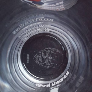 Puede incluir: Un vaso de vidrio transparente con una imagen en blanco y negro de la cara de un hombre con gafas de sol. El vaso tiene las palabras "Iron Maiden" y "Creado por" impresas.