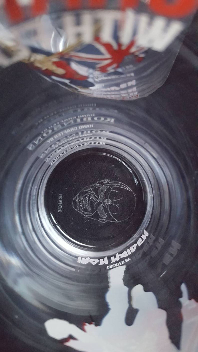 Puede incluir: Un vaso de vidrio transparente con una imagen en blanco y negro de la cara de un hombre con gafas de sol. El vaso tiene las palabras "Iron Maiden" y "Creado por" impresas.