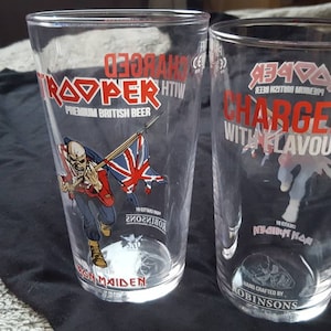 Puede incluir: Dos vasos de pinta de vidrio transparente con el logotipo de la cerveza Iron Maiden Trooper. El vaso de la izquierda tiene una imagen de Eddie, la mascota de Iron Maiden, sosteniendo una bandera de la Union Jack en rojo, blanco y azul. El vaso de la derecha tiene el texto "Charged with Flavour" en rojo y blanco.