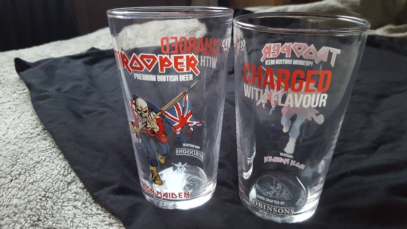 Puede incluir: Dos vasos de pinta de vidrio transparente con el logotipo de la cerveza Iron Maiden Trooper. El vaso de la izquierda tiene una imagen de Eddie, la mascota de Iron Maiden, sosteniendo una bandera de la Union Jack en rojo, blanco y azul. El vaso de la derecha tiene el texto "Charged with Flavour" en rojo y blanco.