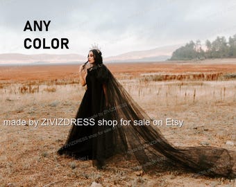 May include: A black tulle wedding dress with a long train and a matching veil. The dress has a fitted bodice and a flowing skirt. The model is wearing a crown and standing in a field.