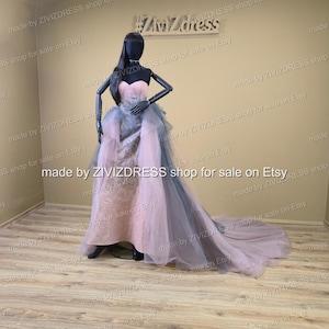 May include: A strapless, floor-length gown on a mannequin. The dress features a pink bodice and a layered skirt with gray and pink tulle. The train extends behind the mannequin. The mannequin is black and has long black hair.
