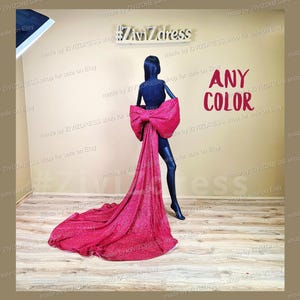 Puede incluir: Una cola de tela roja brillante con un gran lazo en la parte posterior. La cola se muestra en un maniquí. El texto "ANY COLOR" está escrito en rojo en una pared detrás del maniquí. El texto "#ZivZdress" está escrito en negro en un letrero sobre el maniquí.