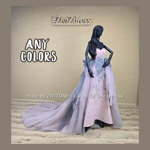 May include: A strapless, floor-length gown in soft pink with a flowing, layered skirt and train. The dress features a gray accent and is displayed on a mannequin. The text "ANY COLORS" is visible.