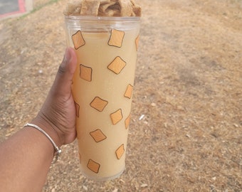 May include: A clear, tall tumbler filled with a light-coloured beverage and topped with cereal. The tumbler is decorated with orange and brown square shapes. The tumbler is held by a person with a silver bracelet.