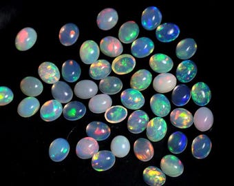 May include: A collection of oval-shaped opals in various colors, including white, blue, green, and pink, are scattered on a black surface. The opals display iridescent flashes of color, creating a shimmering effect. These gemstones are ideal for jewelry making.