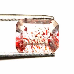 May include: A clear, rectangular gemstone with red inclusions, held by a metal tweezer. The gemstone has a faceted cut, displaying its internal patterns. The inclusions appear as streaks and specks.
