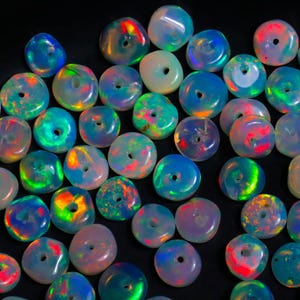 May include: A collection of iridescent opal beads with a hole drilled through the center. The beads display a range of colors, including blue, green, red, and yellow, and are approximately 0.5 inches in diameter. The beads are scattered on a black surface.