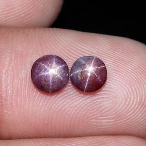 Op de afbeelding: Twee ronde, gepolijste edelstenen met een ster-effect. De edelstenen zijn dieppaars van kleur met een zesstralig sterpatroon in een lichtere tint. De stenen zijn geplaatst tegen een vage achtergrond.