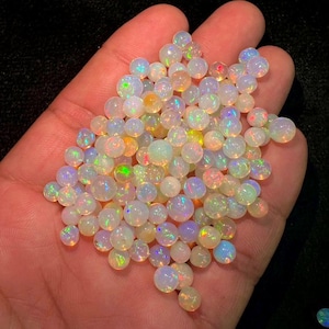 May include: A collection of small, round, translucent white opals featuring a vibrant play-of-colour with flashes of green, orange, and blue. These polished gemstones are spherical in shape and exhibit a smooth, glossy surface texture throughout.