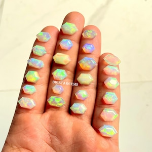May include: A collection of hexagonal opal gemstones in various sizes and colors, including white, yellow, and blue. The gemstones are displayed on a hand, with the text "BIGSTARGEMS" visible.
