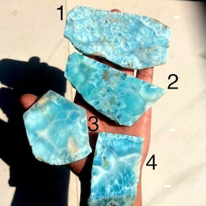 Puede incluir: Cuatro piezas de piedra preciosa de larimar en bruto, cada una con una forma y un patrón únicos. Las piedras muestran una gama de tonos azul claro con vetas blancas, que se asemejan a las olas del océano. Las piedras preciosas se sostienen en una mano.
