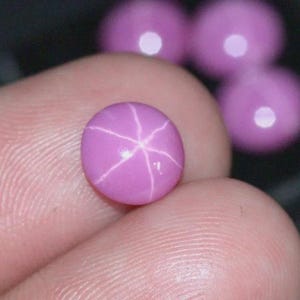 Peut inclure: Gros plan sur une pierre précieuse de rubis étoilé rose. La pierre de forme ovale présente un motif en étoile à six rayons sur sa surface. Plusieurs autres pierres précieuses roses floues sont visibles en arrière-plan. La pierre est tenue entre deux doigts.