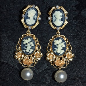 May include: A pair of gold tone earrings with a cameo design. The cameos are white on a black background. The earrings have a floral design and a pearl drop.