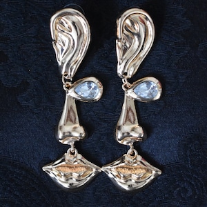 May include: Gold tone earrings with a face design. The earrings feature a teardrop-shaped crystal, a nose, and a pair of lips.