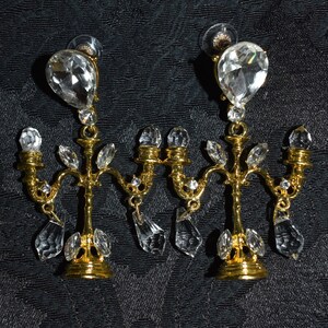 May include: A pair of gold chandelier earrings with clear crystal drops. The earrings are in the shape of a candelabra with five arms.