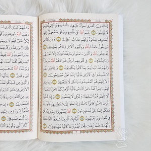 Puede incluir: Libro abierto con escritura &aacute;rabe, con bordes rojos y dorados ornamentados. Las p&aacute;ginas est&aacute;n llenas de texto y el libro descansa sobre una superficie suave, blanca y texturizada. Un texto religioso.