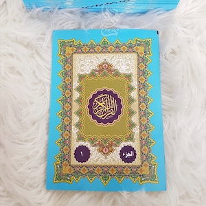 Puede incluir: Una pila de libros azul claro con cubiertas ornamentadas y coloridas. La portada del libro superior presenta dise&ntilde;os dorados, verdes y morados con texto &aacute;rabe. El libro est&aacute; etiquetado con el n&uacute;mero 1 y texto &aacute;rabe.