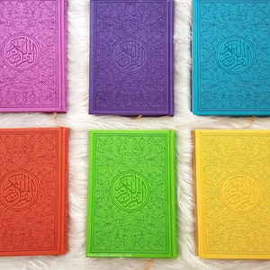 May include: A collection of colorful Quran books with intricate designs and gold accents. The books are arranged in a row on a white fluffy surface.