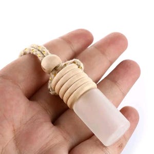 May include: A rectangular, frosted glass car air freshener with a wooden top and a beige rope for hanging. The wooden top has a series of circular ridges. The air freshener is held in a hand against a white background.