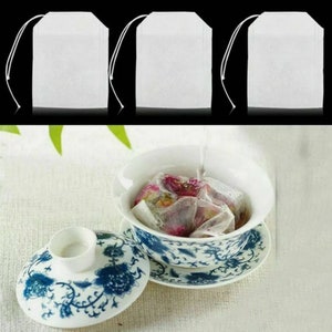 May include: Three white tea bags, made of fine mesh fabric, are shown against a black background. The tea bags are empty, but one is shown in a white porcelain teacup with a blue and white floral design, filled with water and loose tea leaves.
