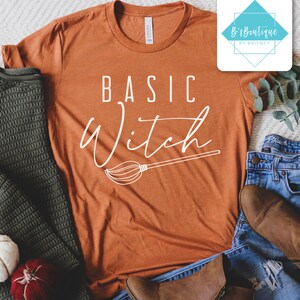 May include: An orange t-shirt with the words "BASIC Witch" in white script and a broom graphic. The shirt is displayed with a green sweater, blue jeans, and brown boots. A small logo is in the upper right corner.