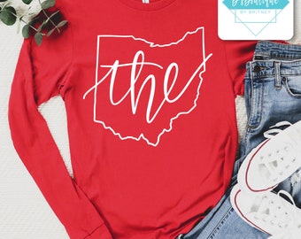 May include: A red long-sleeve shirt featuring a white outline of the state of Ohio with the word "the" in a cursive font. The shirt is paired with blue jeans and white sneakers.
