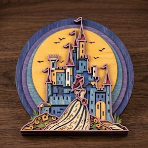 Puede incluir: Una pieza de arte mural de madera multicapa que presenta la silueta de un castillo contra una gran luna amarilla. El diseño incluye una figura con vestido en primer plano, con tonos azules, morados y amarillos, y pequeñas aves negras.