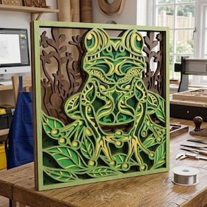 May include: A layered wooden frog art piece. The frog is depicted in shades of green and yellow, with intricate details. The artwork is framed in a green border and set against a brown background with a leafy design. The piece is displayed on a wooden table.