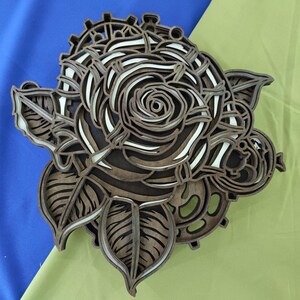 Puede incluir: Una pieza de arte mural de madera multicapa que presenta un diseño de rosa central con intrincados recortes de pétalos. Las capas de madera marrón oscuro están apiladas para crear profundidad, revelando acentos blancos entre las capas.