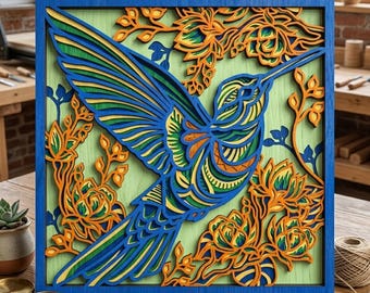 May include: A layered wooden art piece featuring a hummingbird in flight. The artwork showcases a vibrant color palette of blue, green, and orange, set against a light green background within a blue frame. Floral elements surround the bird.