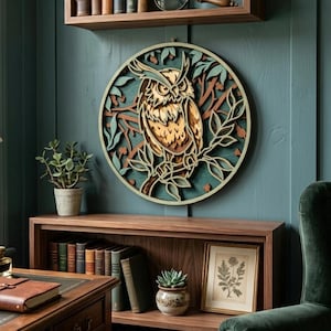 Peut inclure: Une décoration murale ronde en bois multicouche représentant un hibou. Le hibou est perché sur une branche, entouré de feuilles et de branches, sur un fond bleu-vert. Le hibou est brun, beige et crème. L'œuvre est accrochée à un mur bleu-vert.