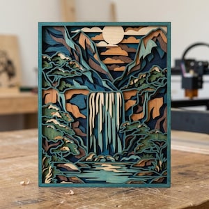 Può includere: Un'opera d'arte in legno a strati raffigurante una scena di cascata. L'opera presenta una cascata che scende da una montagna, con alberi e un lago alla base. La tavolozza dei colori comprende sfumature di blu, verde, marrone e beige, con un sole chiaro nel cielo.
