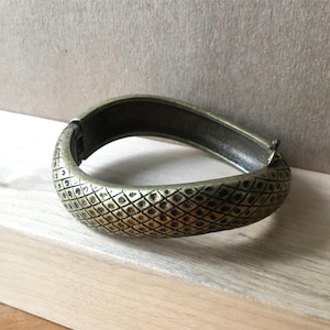 May include: A hinged, antique gold-tone bracelet with a textured, diamond-patterned surface. The bracelet is open, revealing the inner surface and clasp. The jewelry is displayed on a light-colored wooden surface.