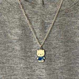 May include: A silver chain necklace with a Hello Kitty pendant. The pendant features a cream-colored cat with a blue outfit. The necklace is displayed on a gray t-shirt.