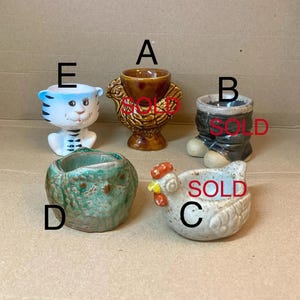 Può includere: Cinque porta uova in ceramica con diversi disegni di animali. Un pollo marrone, un pollo grigio e bianco, un gufo verde, una tigre blu e bianca e un uomo in jeans grigi e neri.