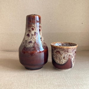 May include: A vintage ceramic salt shaker and matching cup. The salt shaker is a tall, glossy brown with a speckled tan pattern. The cup has a similar design. Both pieces have a mid-century modern aesthetic.
