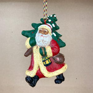 May include: A ceramic Christmas ornament depicting Santa Claus carrying a green Christmas tree. Santa is wearing a red suit with a white trim, a white beard, and a black hat. He is carrying a lantern and a bag.