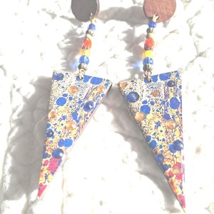 Peut inclure: Une paire de boucles d'oreilles uniques avec un dessus en disque de bois et un pendentif triangulaire coloré. Les pendentifs ont un motif moucheté avec des accents bleus, jaunes et rouges. Les boucles d'oreilles sont reliées par un fil de perles colorées.