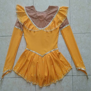 K&ouml;nnte beinhalten: Ein gelbes Eiskunstlaufkleid mit langen &Auml;rmeln und einem R&uuml;schenkragen. Das Kleid ist mit Strasssteinen und einem silbernen Besatz verziert.