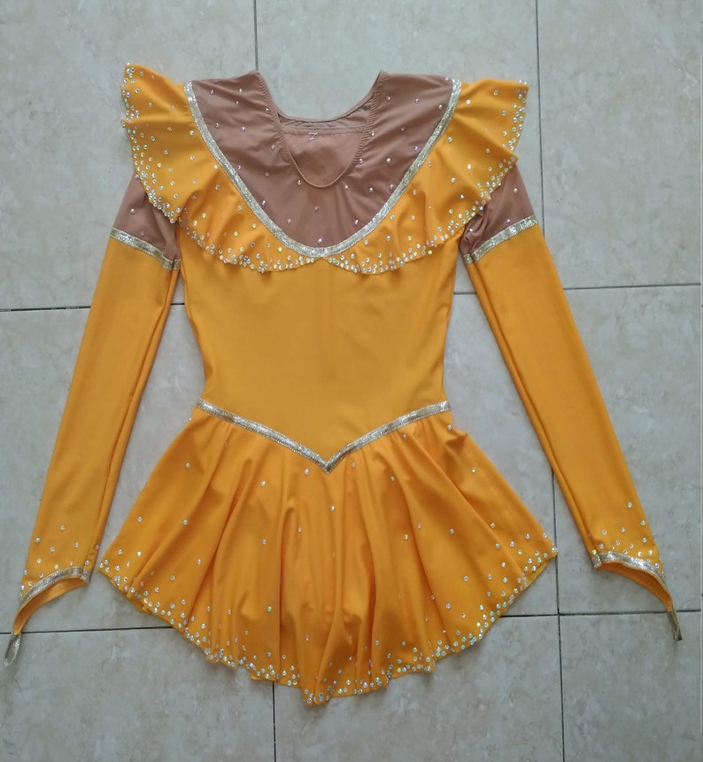 K&ouml;nnte beinhalten: Ein gelbes Eiskunstlaufkleid mit langen &Auml;rmeln und einem R&uuml;schenkragen. Das Kleid ist mit Strasssteinen und einem silbernen Besatz verziert.