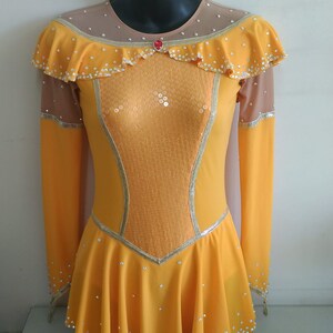 K&ouml;nnte beinhalten: Ein gelbes Eiskunstlaufkleid mit langen &Auml;rmeln und einem R&uuml;schenkragen. Das Kleid ist mit Pailletten und Strasssteinen verziert.