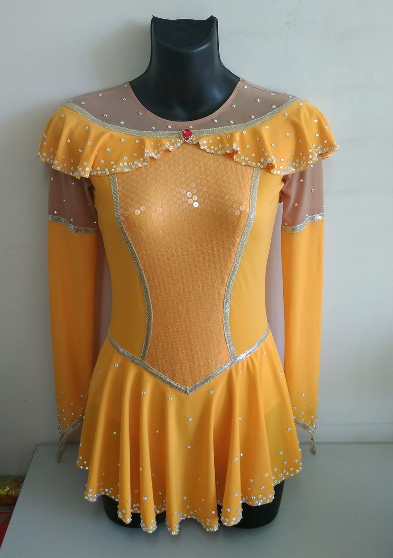 K&ouml;nnte beinhalten: Ein gelbes Eiskunstlaufkleid mit langen &Auml;rmeln und einem R&uuml;schenkragen. Das Kleid ist mit Pailletten und Strasssteinen verziert.