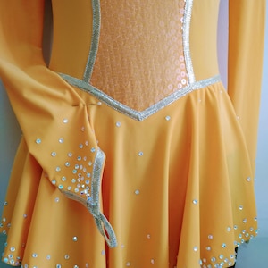 K&ouml;nnte beinhalten: Ein gelbes Eiskunstlaufkleid mit langen &Auml;rmeln und einem Paillettenoberteil. Das Kleid ist mit Strasssteinen am Saum und an den &Auml;rmeln verziert.