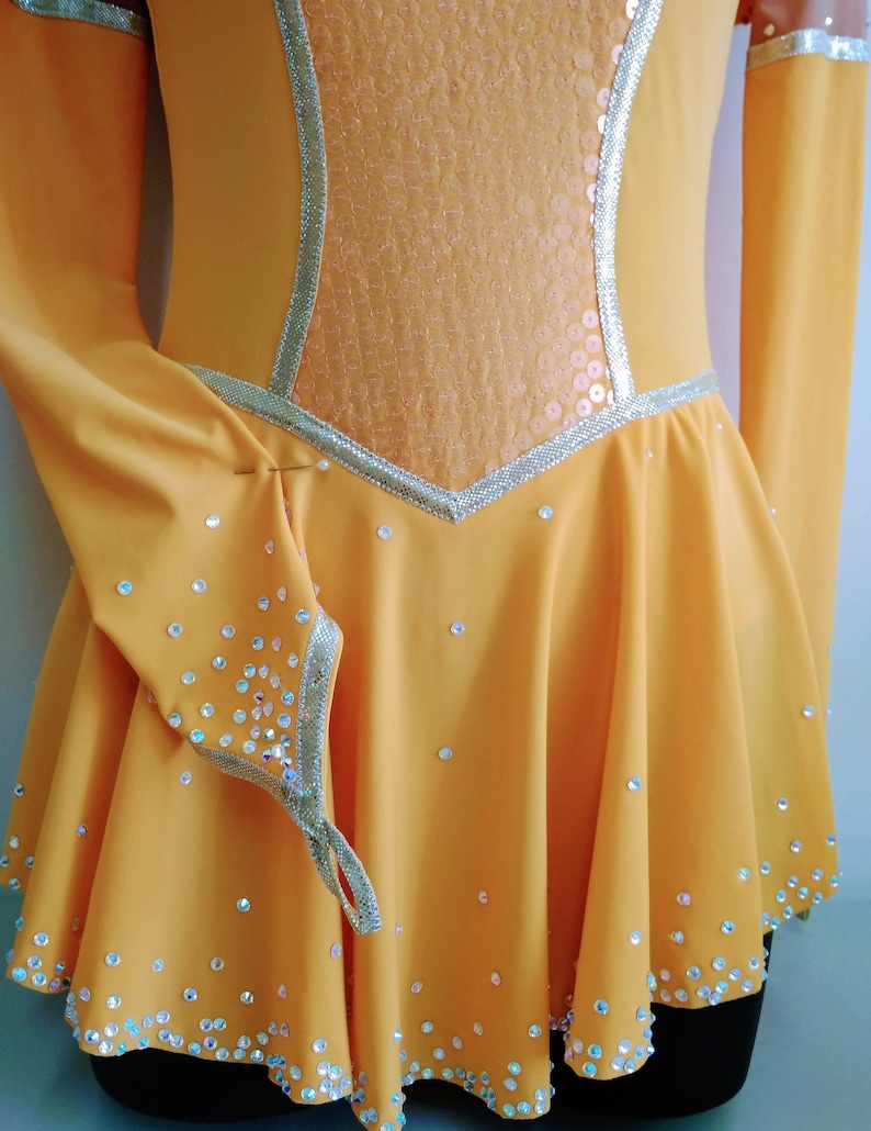 K&ouml;nnte beinhalten: Ein gelbes Eiskunstlaufkleid mit langen &Auml;rmeln und einem Paillettenoberteil. Das Kleid ist mit Strasssteinen am Saum und an den &Auml;rmeln verziert.