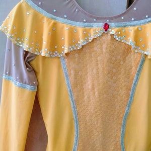 K&ouml;nnte beinhalten: Ein gelbes und braunes Eiskunstlaufkleid mit einem R&uuml;schenkragen, einem roten Strassstein und einem durchsichtigen Einsatz in der Mitte.